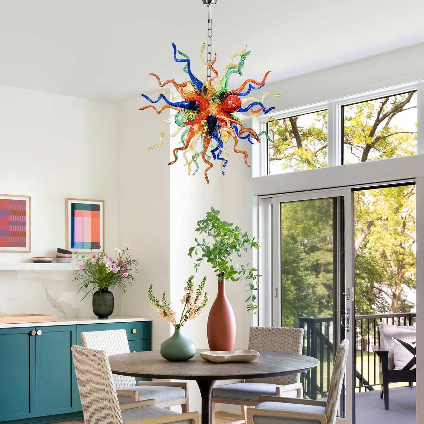 Multi-color hand blown glass chandelier brightly lit against a white wall with a large window, showcasing its radiant glow.