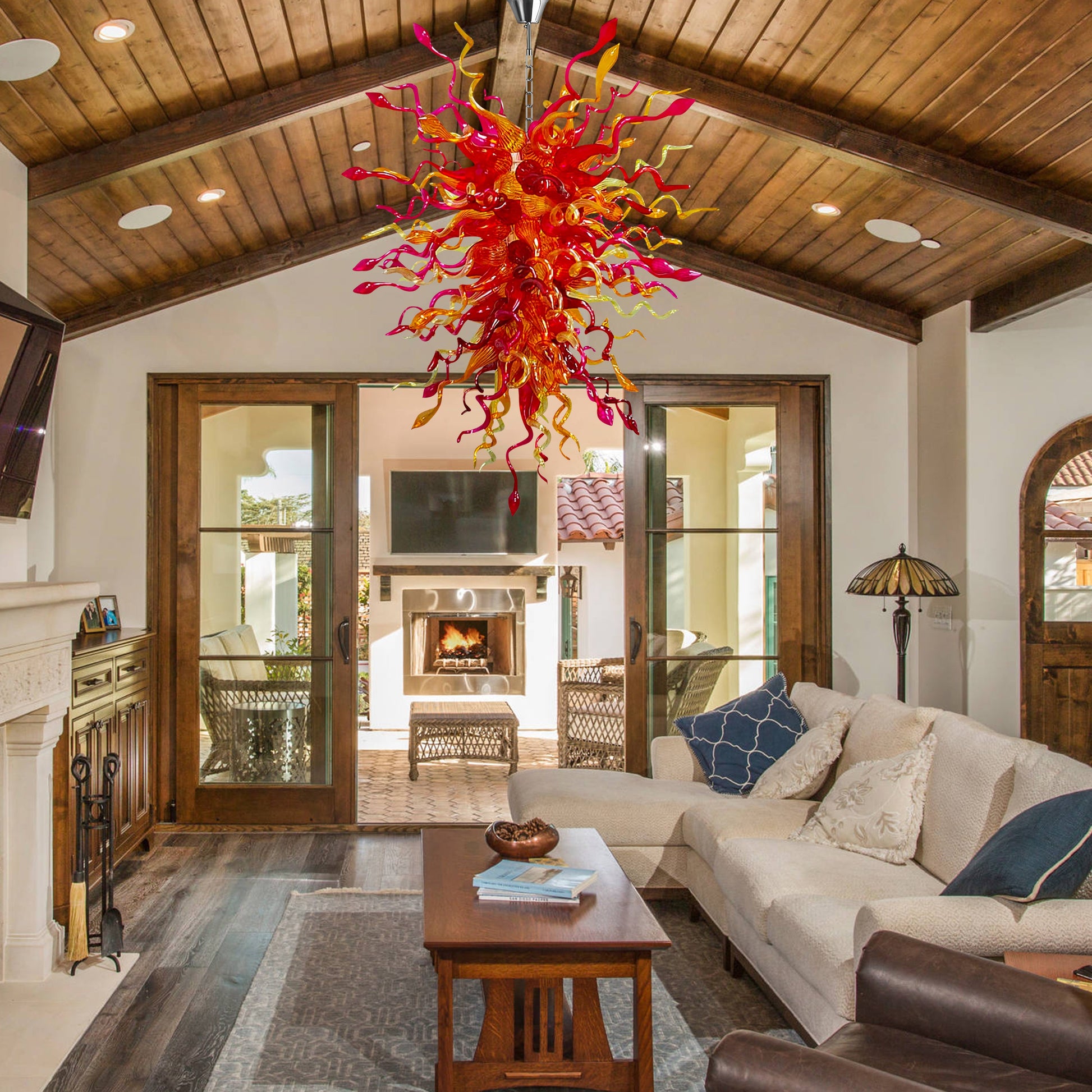 Hand blown glass chandelier with red and yellow hues hanging from a vaulted wooden ceiling in a cozy living room with a fireplace.
