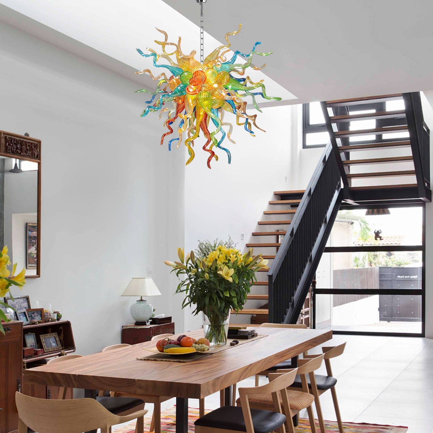Modern dining room with a colorful Chihuly blown glass chandelier, wooden table, and staircase.